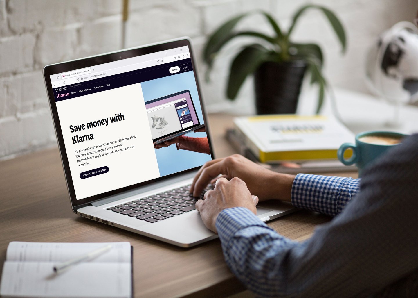 Person using a laptop displaying the Klarna website with a ‘Save money with Klarna’ message, at a desk with a notebook, coffee mug, and books