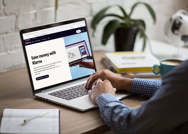 Person using a laptop displaying the Klarna website with a ‘Save money with Klarna’ message, at a desk with a notebook, coffee mug, and books